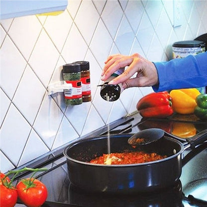 Adhesive spice rack mounted on a kitchen wall holding two spice jars near a stovetop while cooking