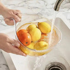 Hands rinsing lemons and an orange in the clear colander bowl under a kitchen faucet